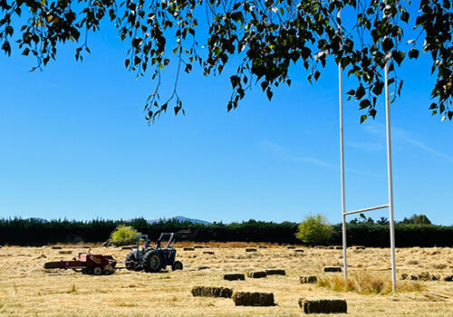 A square hay baler picks up bales in a dry field with a large blue sky above. The field contains a full sized rugby goal.
