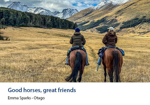 Two riders on horseback make their way through a wide alpine valley surrounded by snow-capped hills. Caption: Emma Sparks, Otago.