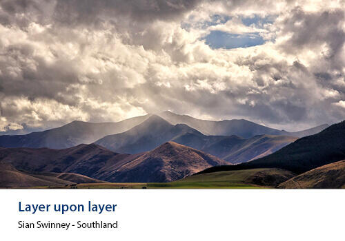 Overlapping ridges and mountains create depth under a cloudy sky with dramatic light falling across the Southland landscape. Caption: Sian Swinney, Southland.