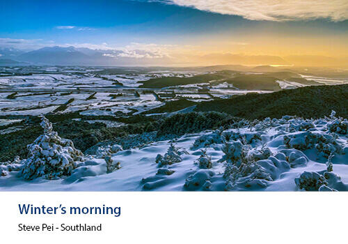 A snow-covered landscape glows in blue light, with sunrise casting a golden hue across distant hills. Caption: Steve Pei, Southland.