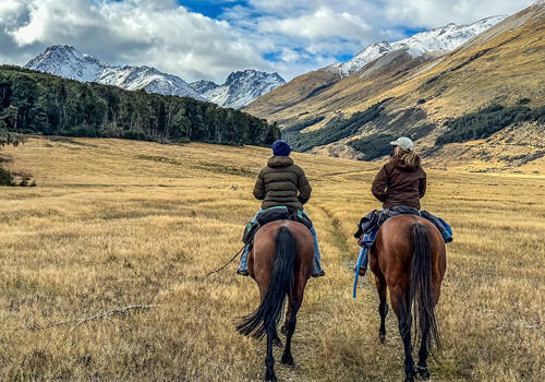 Two riders on horseback make their way through a wide alpine valley surrounded by snow-capped hills. Caption: "Good horses, great friends" Emma Sparks, Otago.