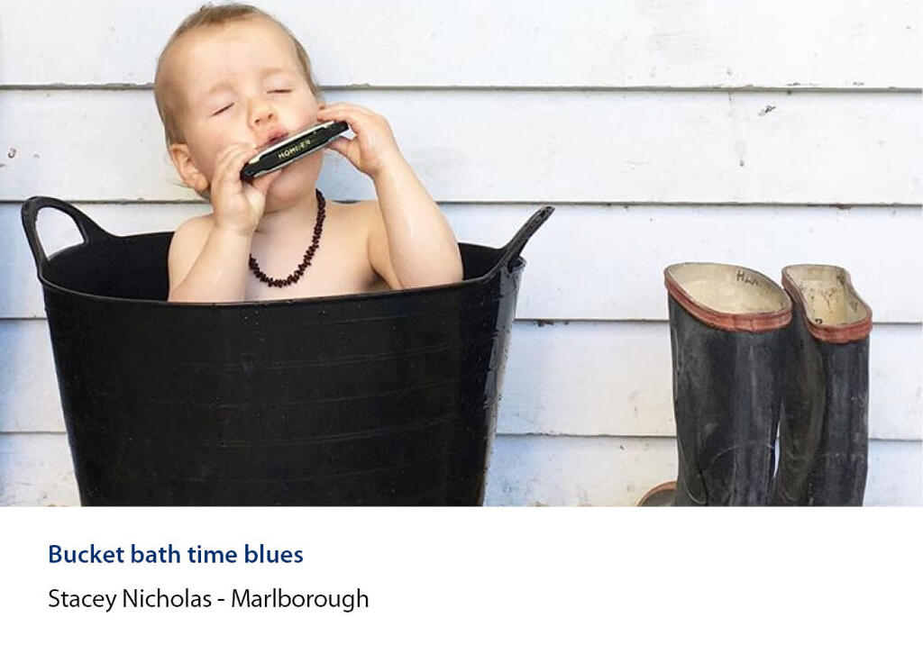 Toddler in a tub bath playing a harmonica beside a pair of red band gumboots.
