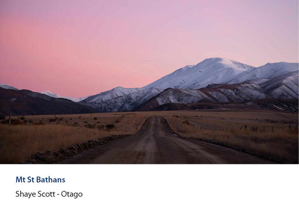 Gravel road running towards snow-covered Mt St Bathans under a gradient pink dusk sky.